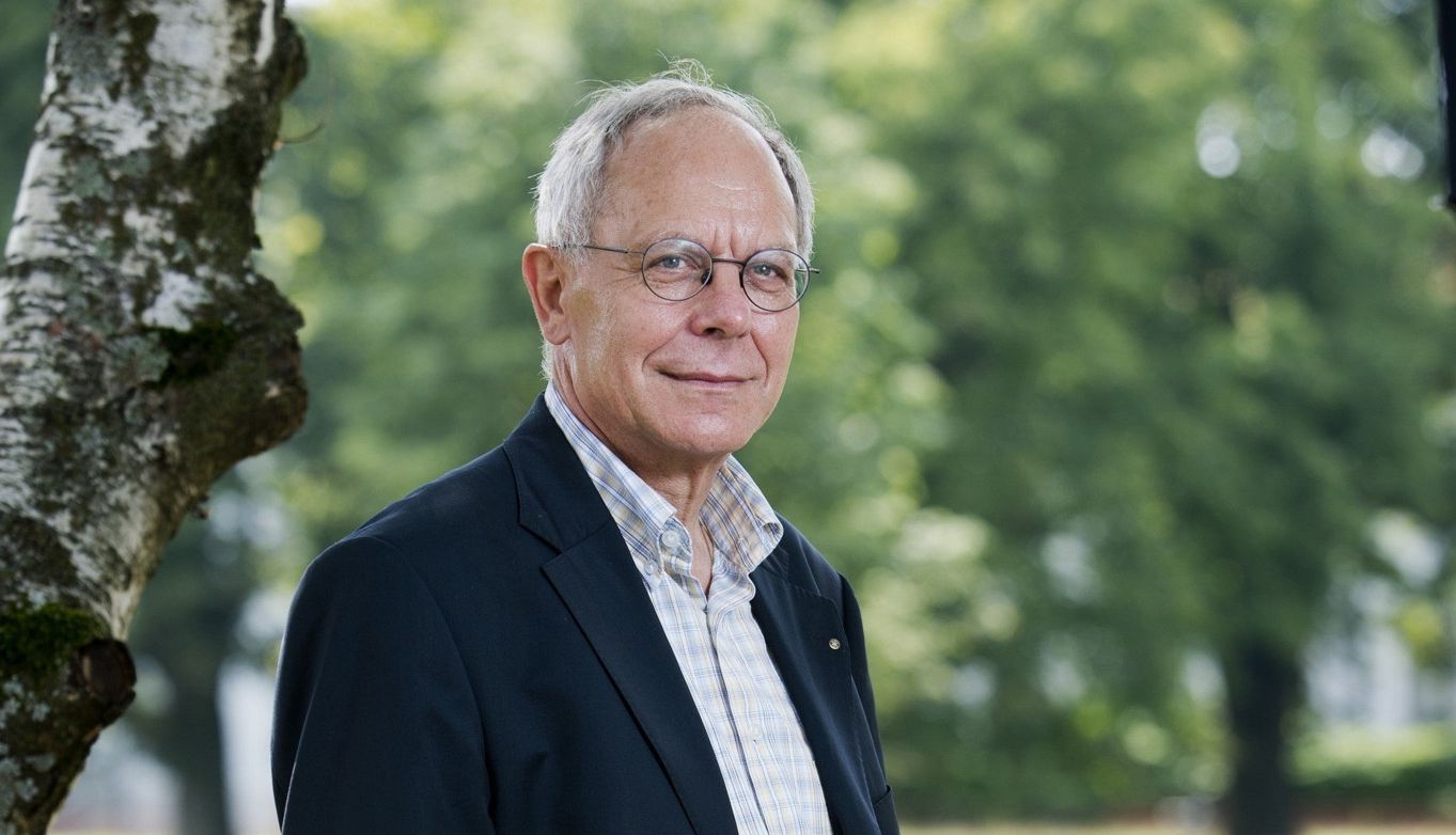 Un homme âgé en costume décontracté se tient debout dans un parc, avec des arbres en arrière-plan.