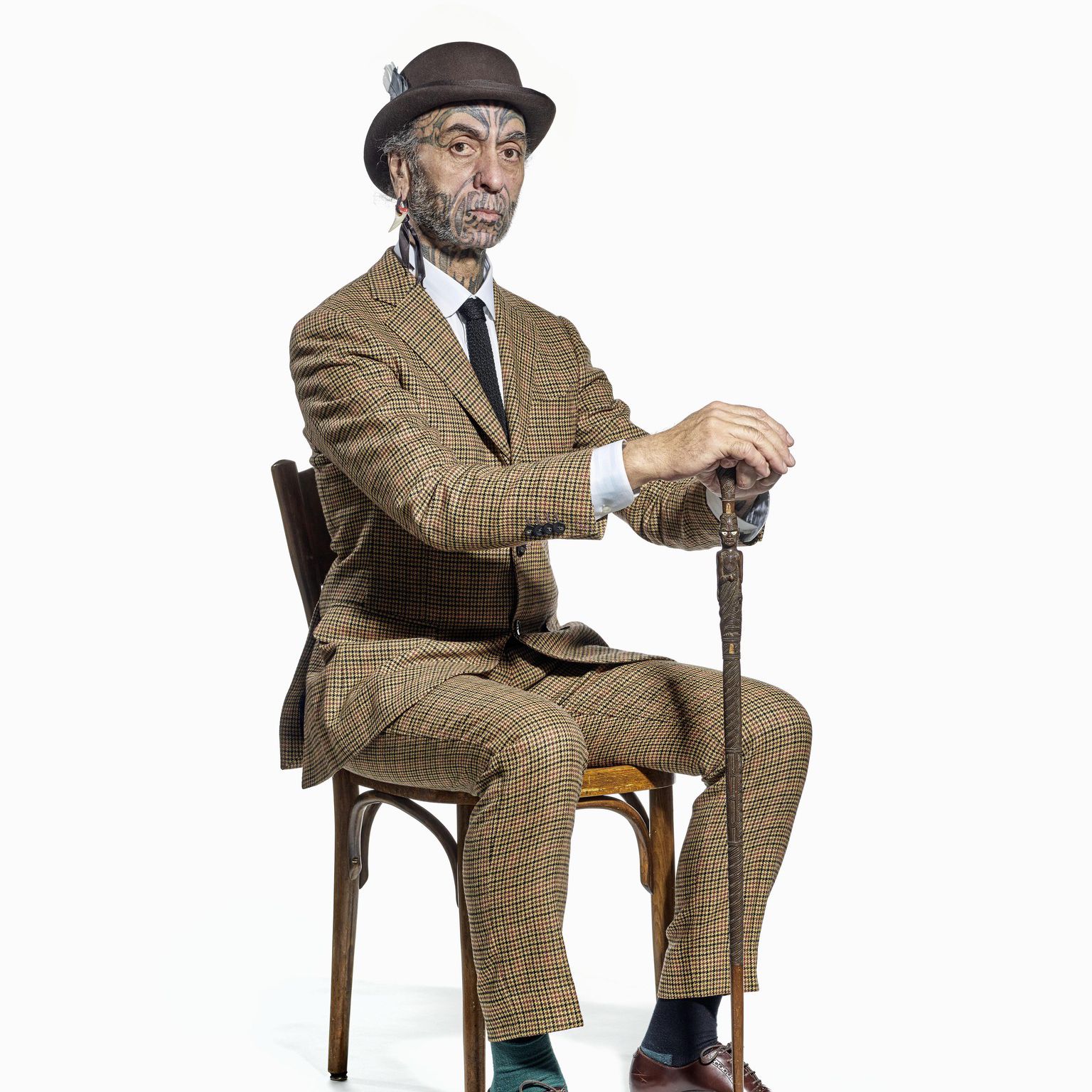 Homme assis sur une chaise, vêtu d’un costume marron, avec un chapeau et une canne.