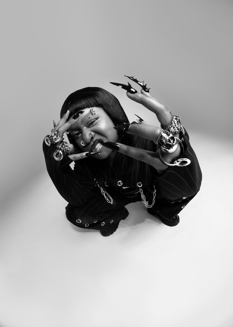 Personne posant en noir et blanc, habillée de vêtements sombres avec de nombreux accessoires et des ongles extravagants.