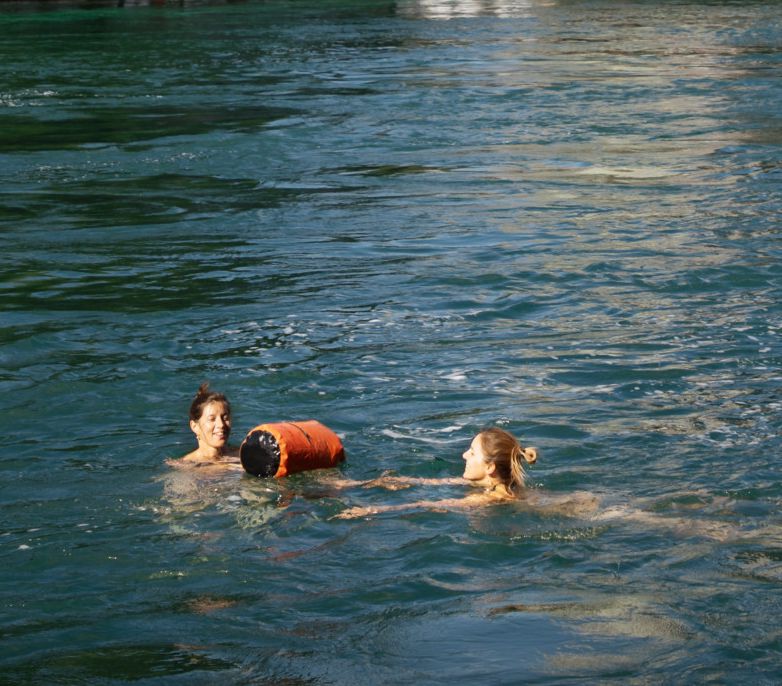Deux personnes nageant dans une rivière avec un pont et un immeuble en arrière-plan, par une journée ensoleillée.