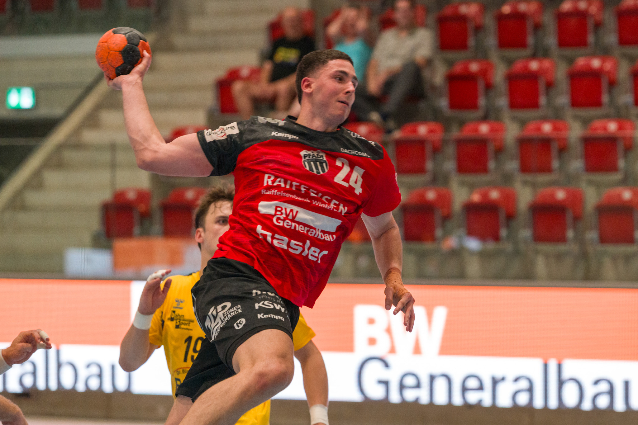 Handballer mit Nummer 24 im rot-schwarzen Trikot springt zum Wurf mit orangem Ball in einer Sporthalle.