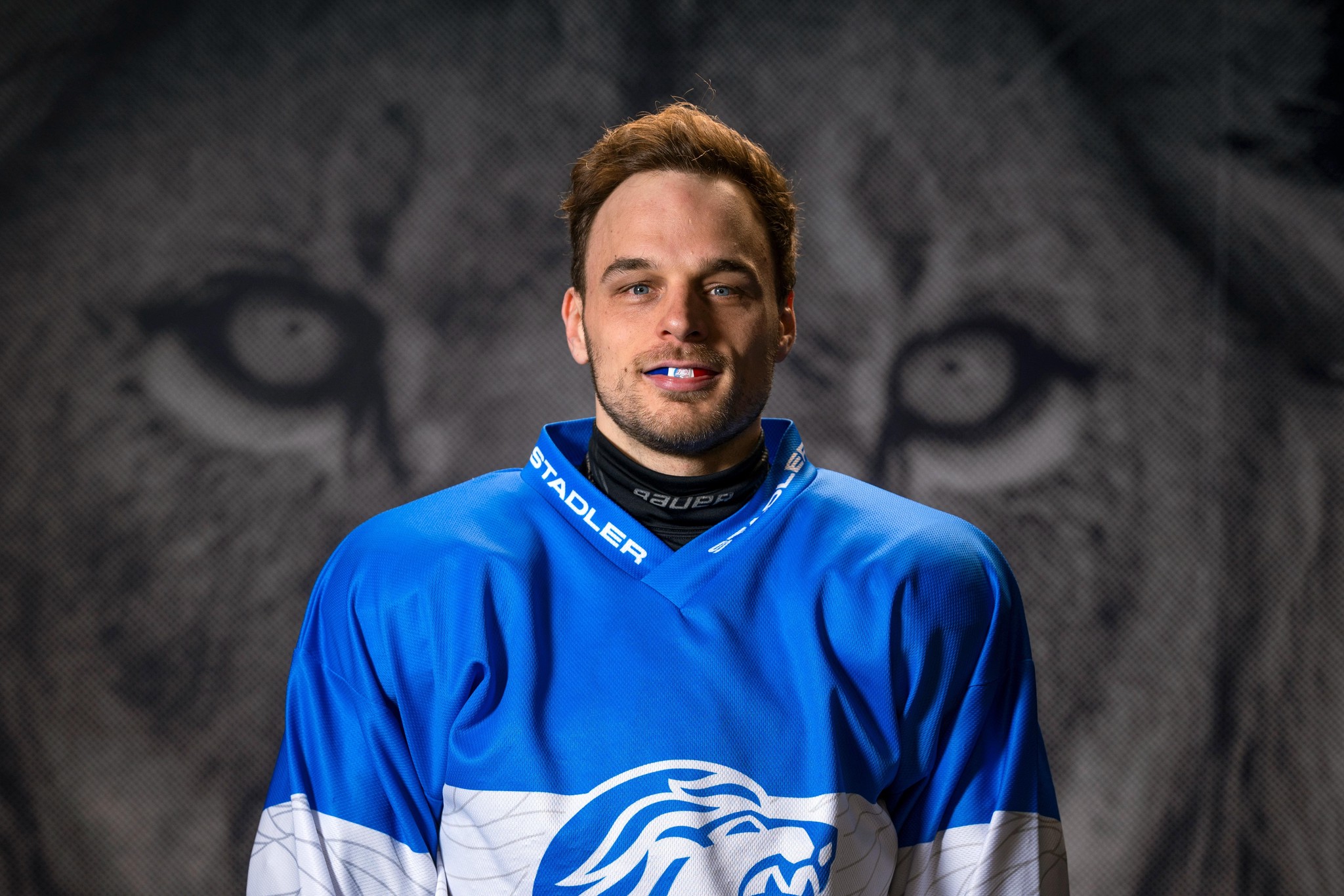 Eishockeyspieler Chris Baltisberger in blauem Trikot mit Löwenemblem, Hintergrund zeigt das Gesicht eines Löwen.