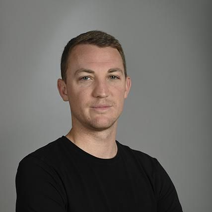 Un homme portant un t-shirt noir, les bras croisés, se tient devant un fond gris uni.