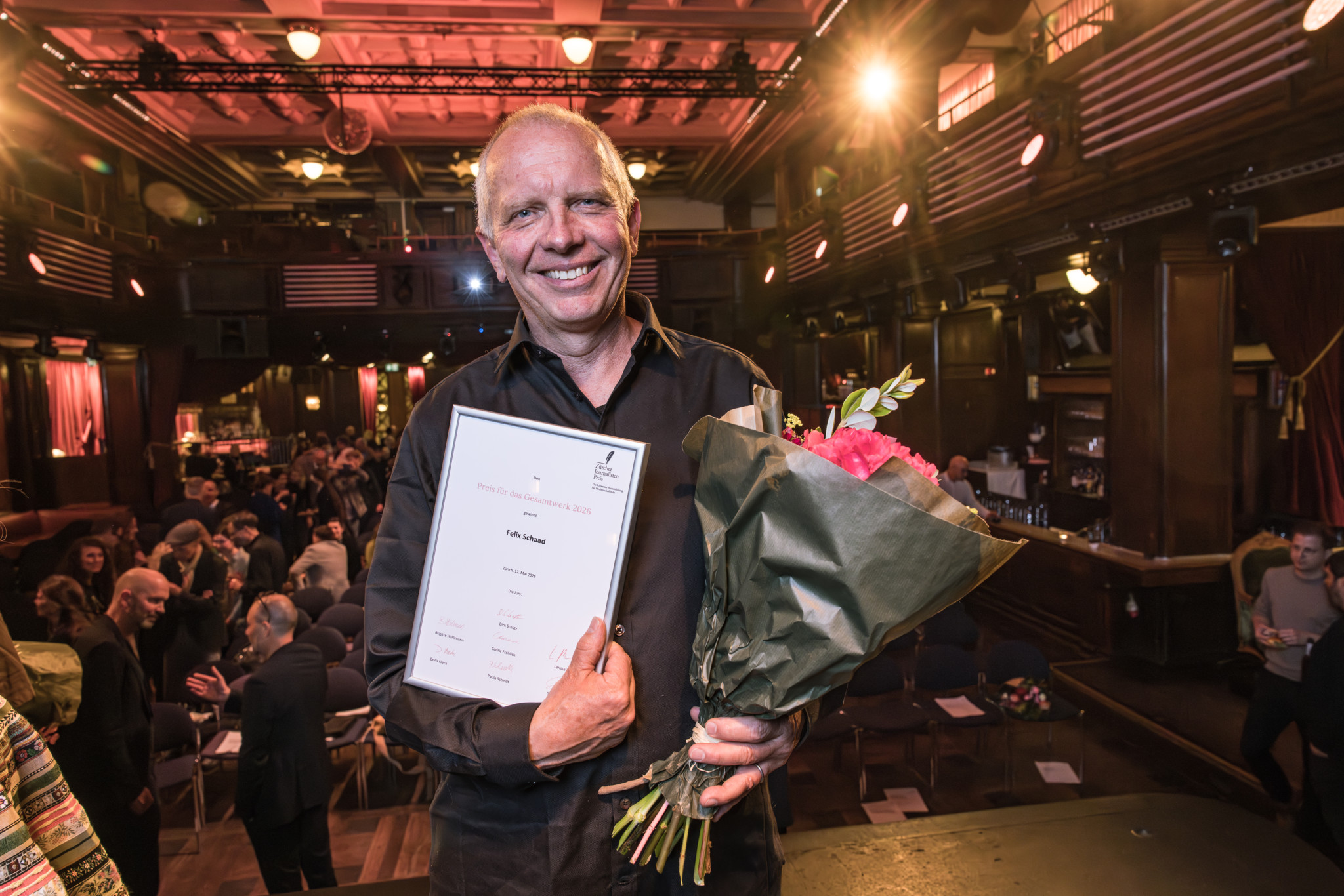 Ein Mann lächelt stolz und hält eine Auszeichnung sowie einen Blumenstrauss an der Verleihung des 46. Zürcher Journalistenpreises.