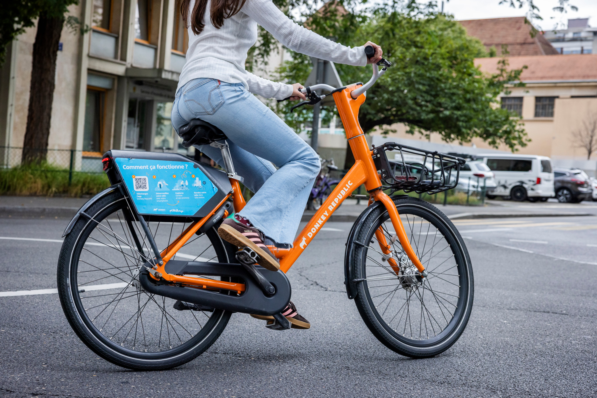 Une personne en jean et pull blanc utilise un vélo orange Donkey Republic en libre-service à Genève.