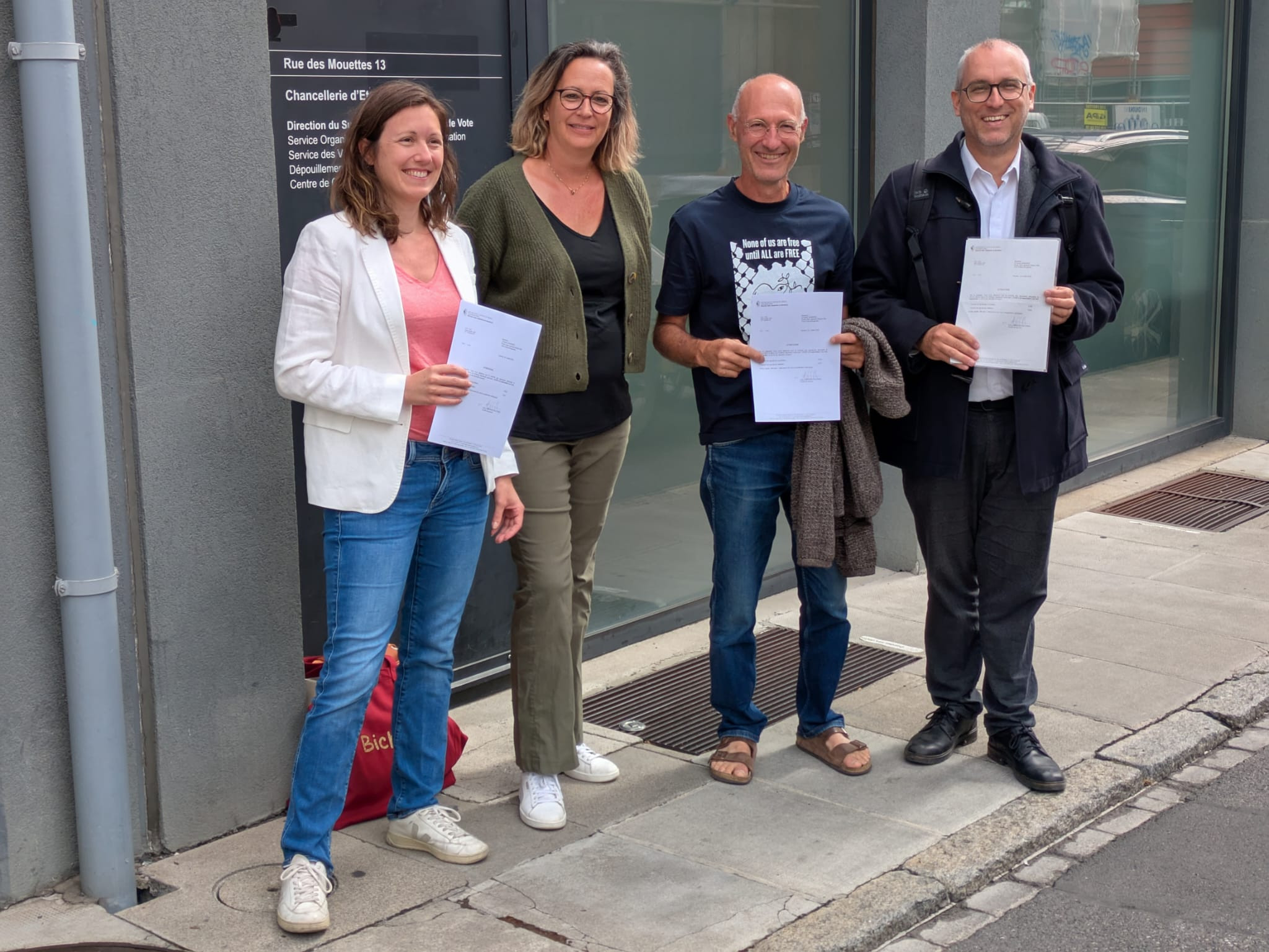 Quatre personnes souriantes tenant des documents, debout devant un bâtiment avec une plaque.
