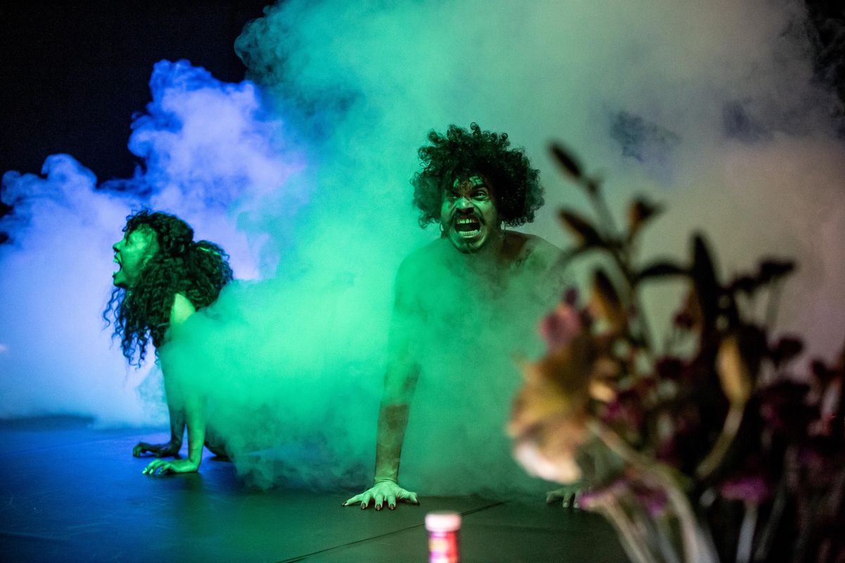 Deux performeurs avec des expressions faciales intenses dans une scène enveloppée de fumée colorée, éclairée par des lumières vertes.