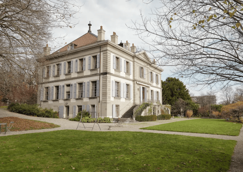 Grande maison blanche entourée d’arbres et de jardin en automne, ciel nuageux.