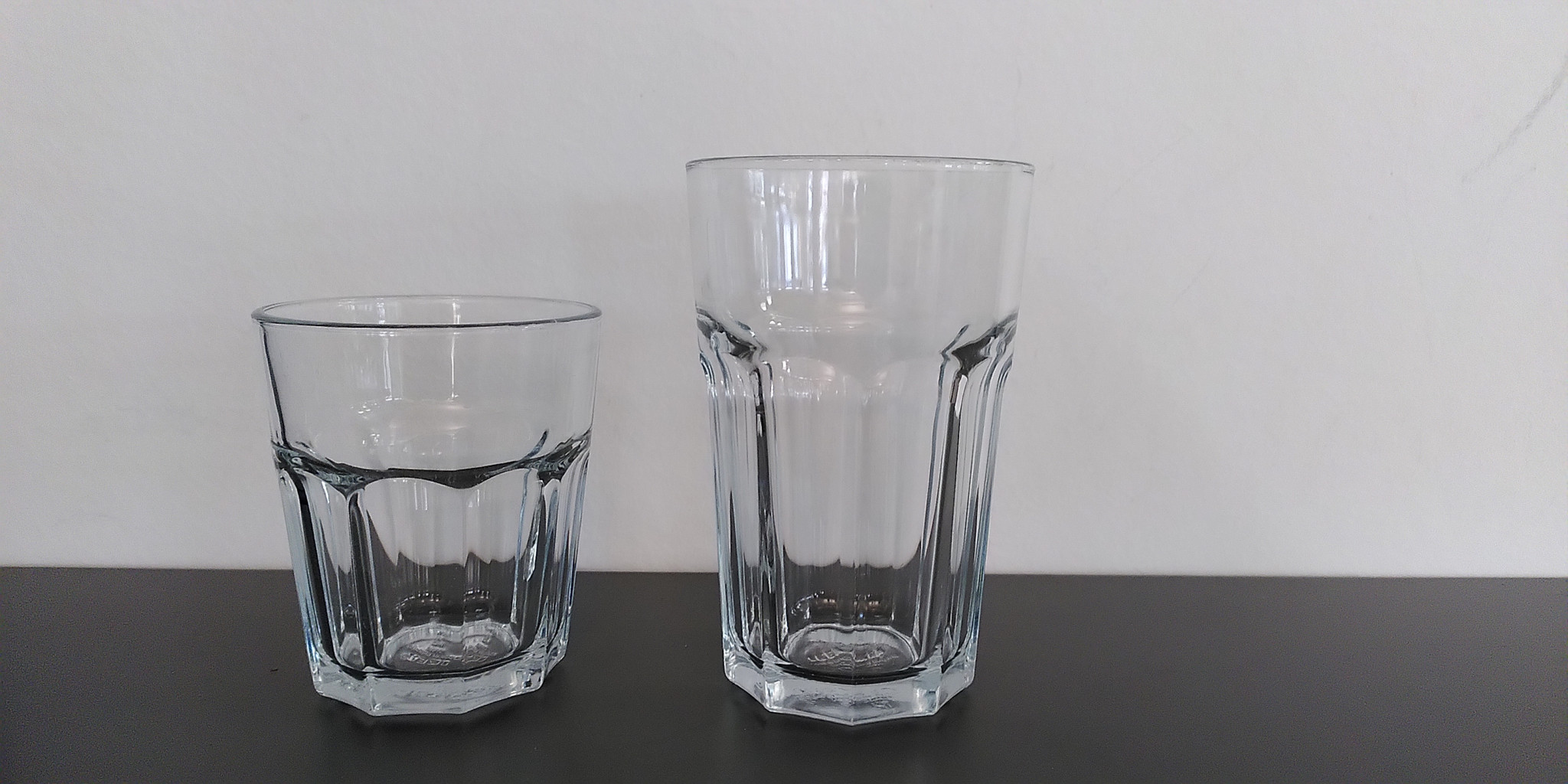 Zwei leere Trinkgläser aus Glas, ein kleineres und ein grösseres, stehen nebeneinander auf einer dunklen Oberfläche vor einer weissen Wand.