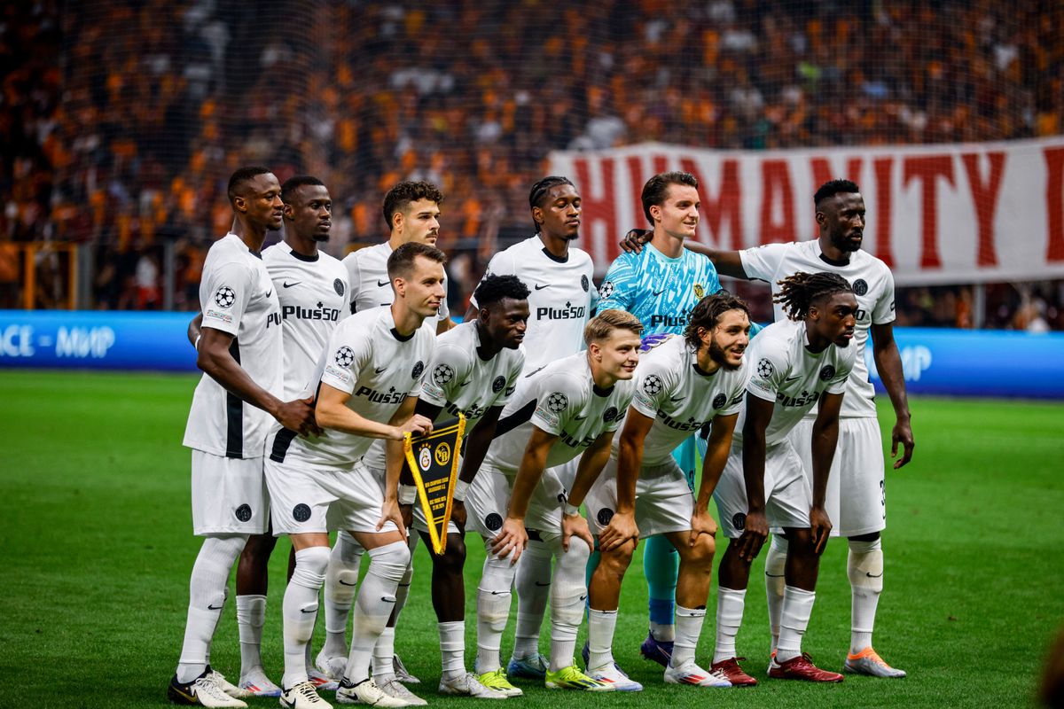 Équipe de football posant avant un match, joueurs en maillots blancs avec écussons noirs et logo 'Plus500', certains à genoux, d'autres debout. Supporters et bannière 'HUMANITY R8' en arrière-plan.