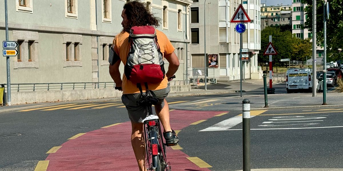 Homme avec un sac à dos rouge et blanc roulant à vélo sur une piste cyclable en milieu urbain.