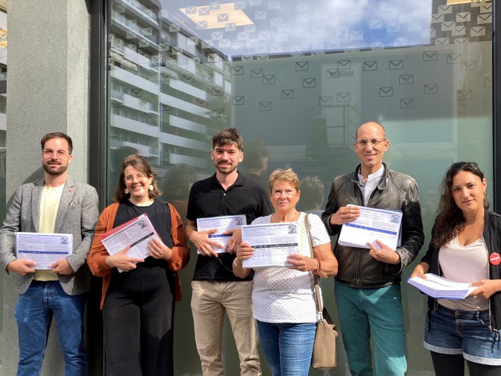 Un groupe de six personnes debout devant un bâtiment, tenant des documents dans leurs mains. Le bâtiment a des fenêtres avec des motifs d'enveloppes.