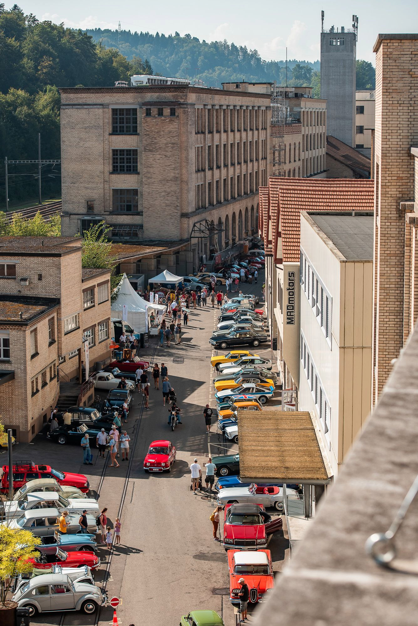 Strassenszene mit zahlreichen geparkten Oldtimern und Menschen, die durch eine historische Industriearchitektur schlendern, im Hintergrund grüne Hügel.