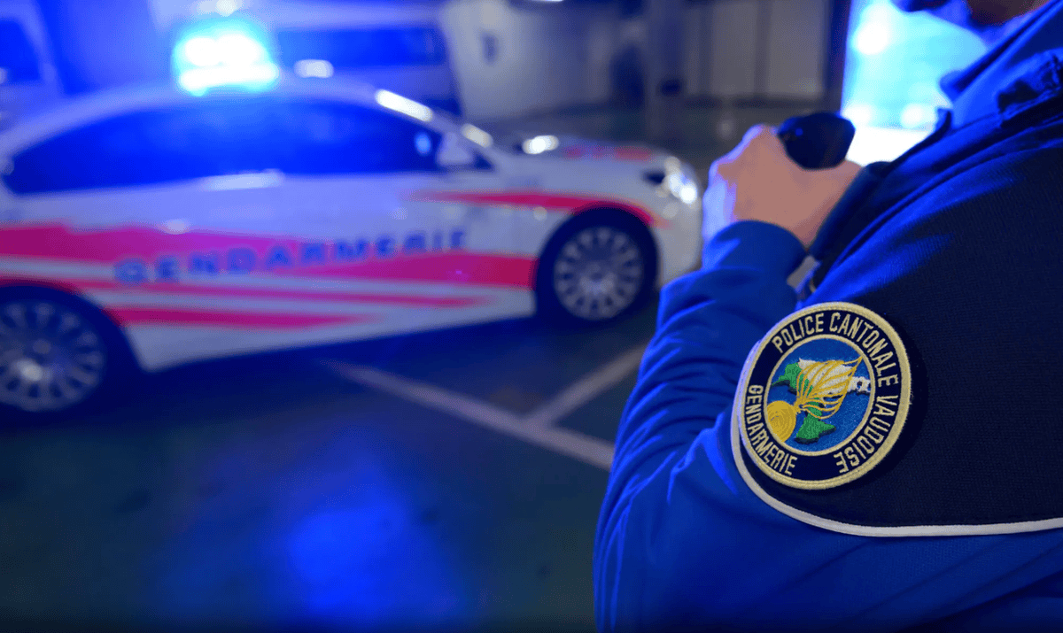 Un policier de la gendarmerie cantonale vaudoise parle à la radio devant une voiture de gendarmerie avec des lumières bleues allumées.