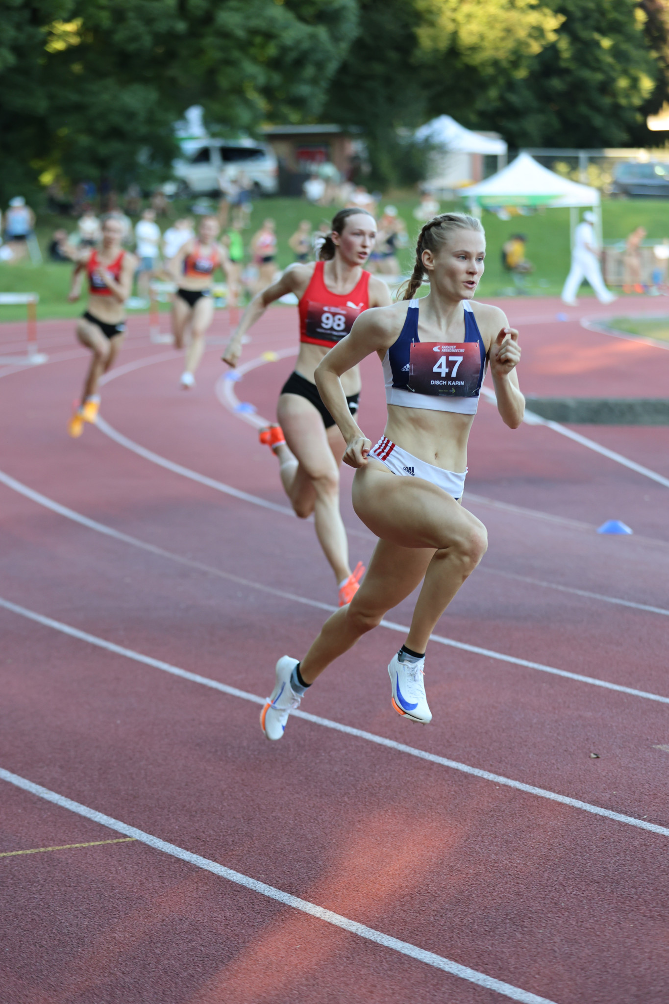 Läuferinnen in einem Wettkampf rennen auf einer Leichtathletikbahn. Eine Läuferin mit der Nummer 47 führt das Rennen vor anderen Konkurrenzteilnehmerinnen.