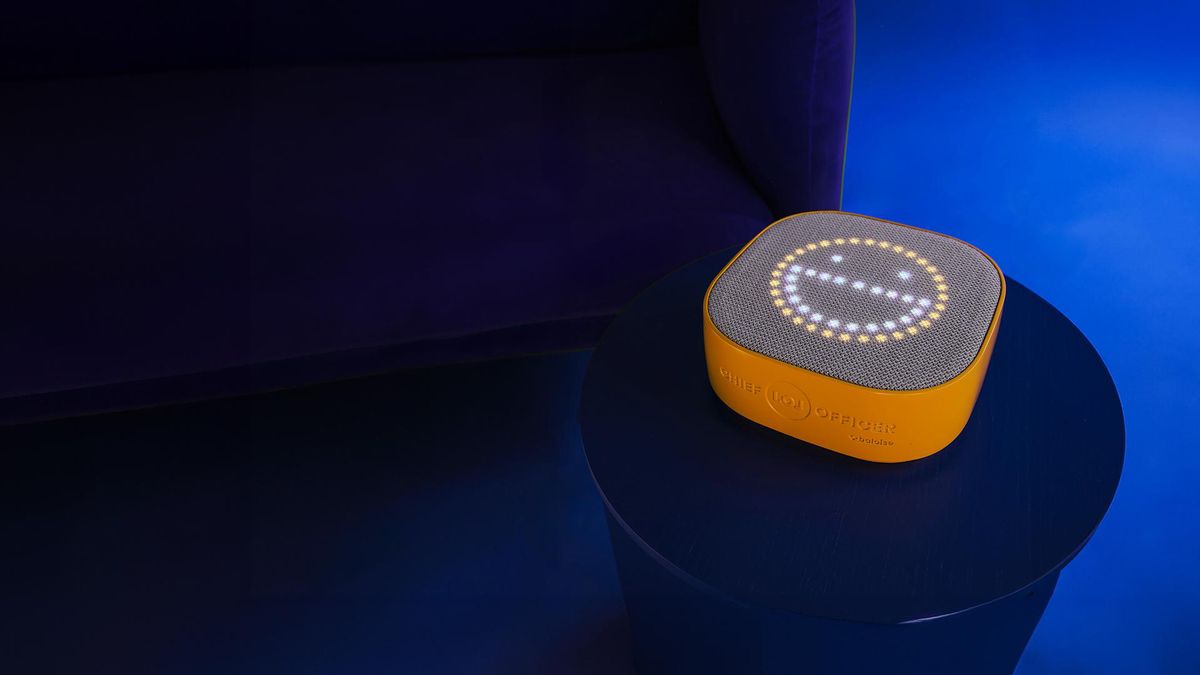 Haut-parleur intelligent jaune et gris, affichant un visage souriant avec des LED blanches, posé sur une table bleue.