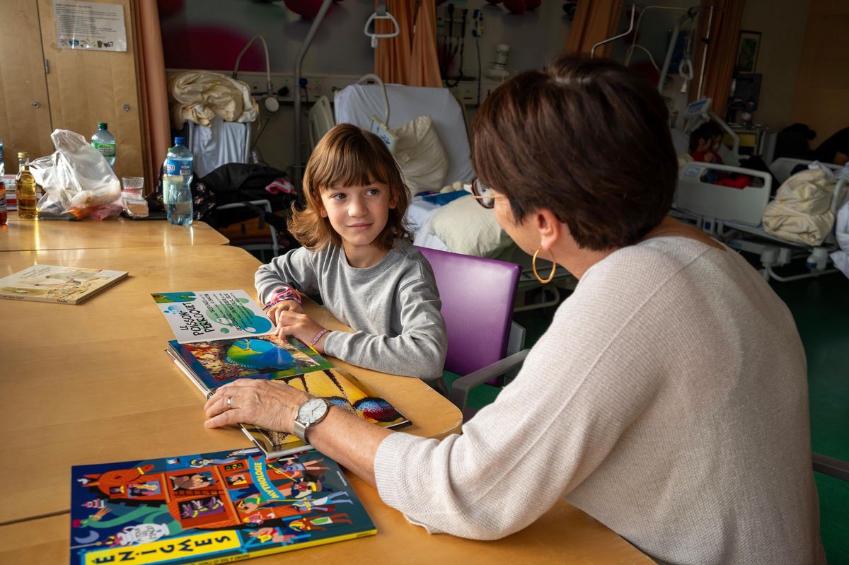 Une enseignante parle avec une jeune élève dans un hôpital, entourées de livres colorés sur une table, illustrant un programme scolaire pour enfants hospitalisés à Lausanne.