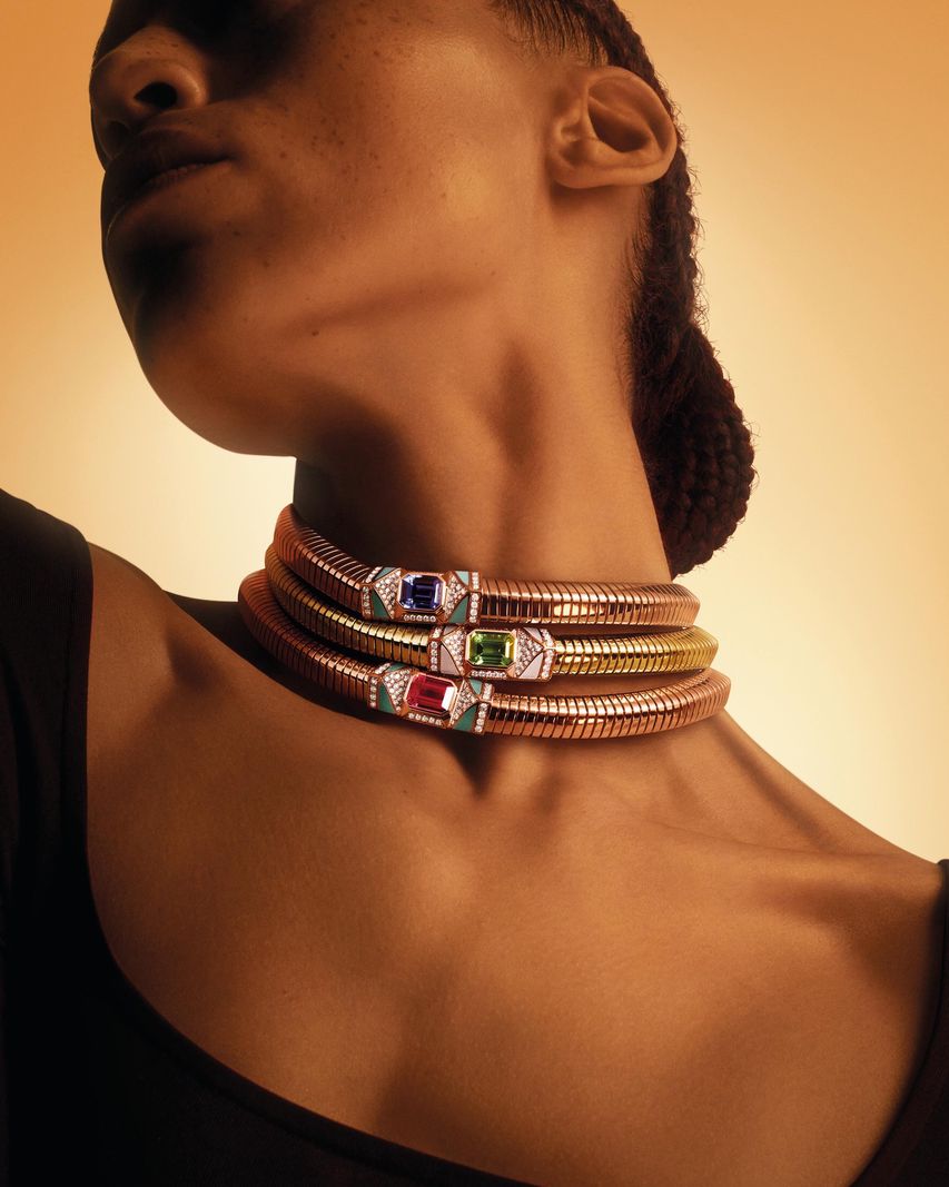 Personne portant plusieurs colliers en or avec des pierres précieuses sur un fond dégradé orange et brun.