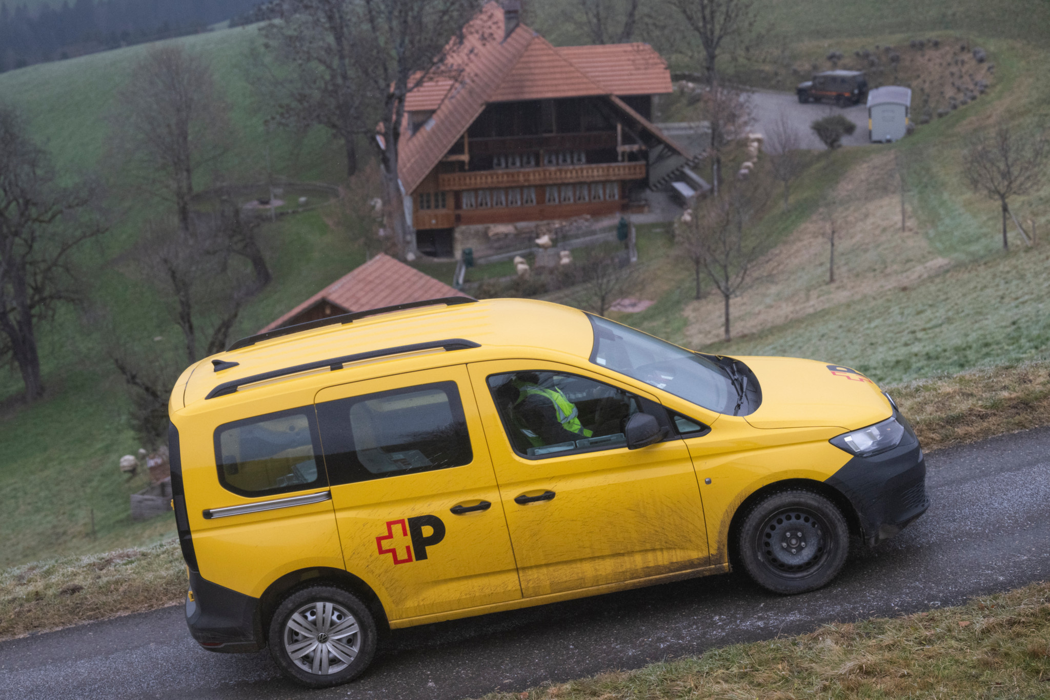 Ein gelbes Schweizer Postauto fährt an einem bewaldeten Berghang in Hinterfultigen entlang. Im Hintergrund ist ein traditionelles Holzhaus zu sehen.