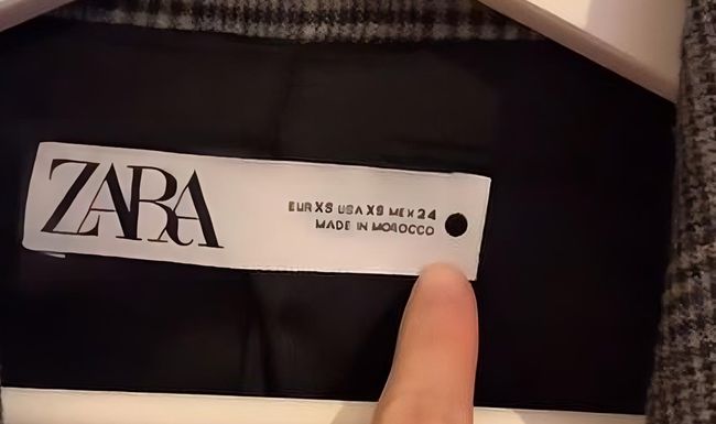 Finger zeigt auf ein rundes Symbol auf der Etikette von einem Kleidungsstück von Zara.