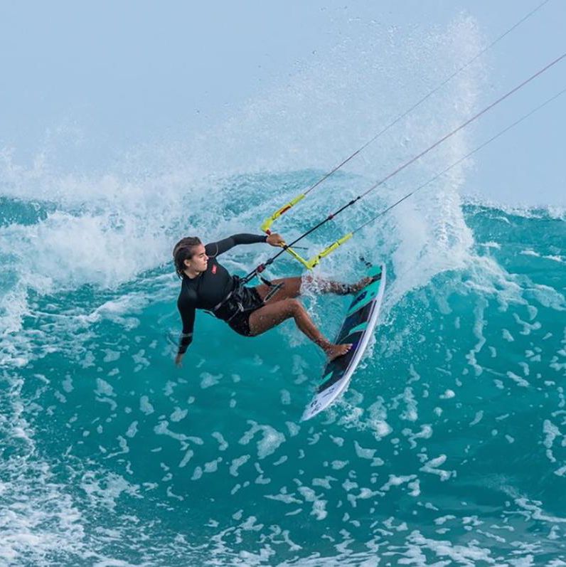 Kitesurfeur effectuant une manœuvre spectaculaire sur une vague turquoise sous un ciel dégagé.