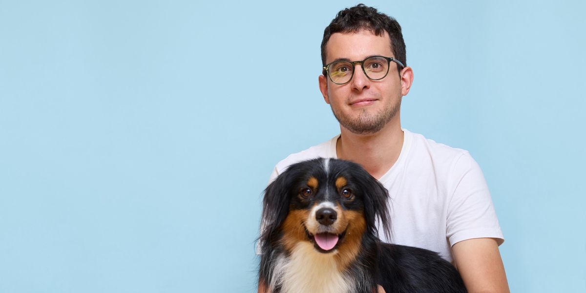 Un homme assis en tailleur avec un chien de race berger australien sur ses genoux, devant un fond bleu.