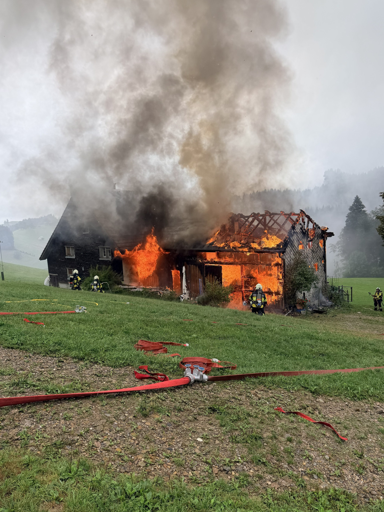 Brennendes Holzhaus auf einer Wiese mit Feuerwehrleuten im Einsatz, dichtem Rauch und roten Feuerwehrschläuchen im Vordergrund.