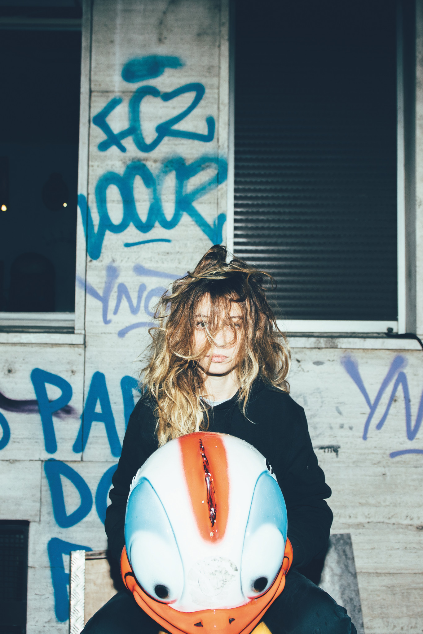 Junge Frau mit langen blonden Haaren hält einen bunten Helm vor einer mit FCZ-Graffiti besprühten Wand.