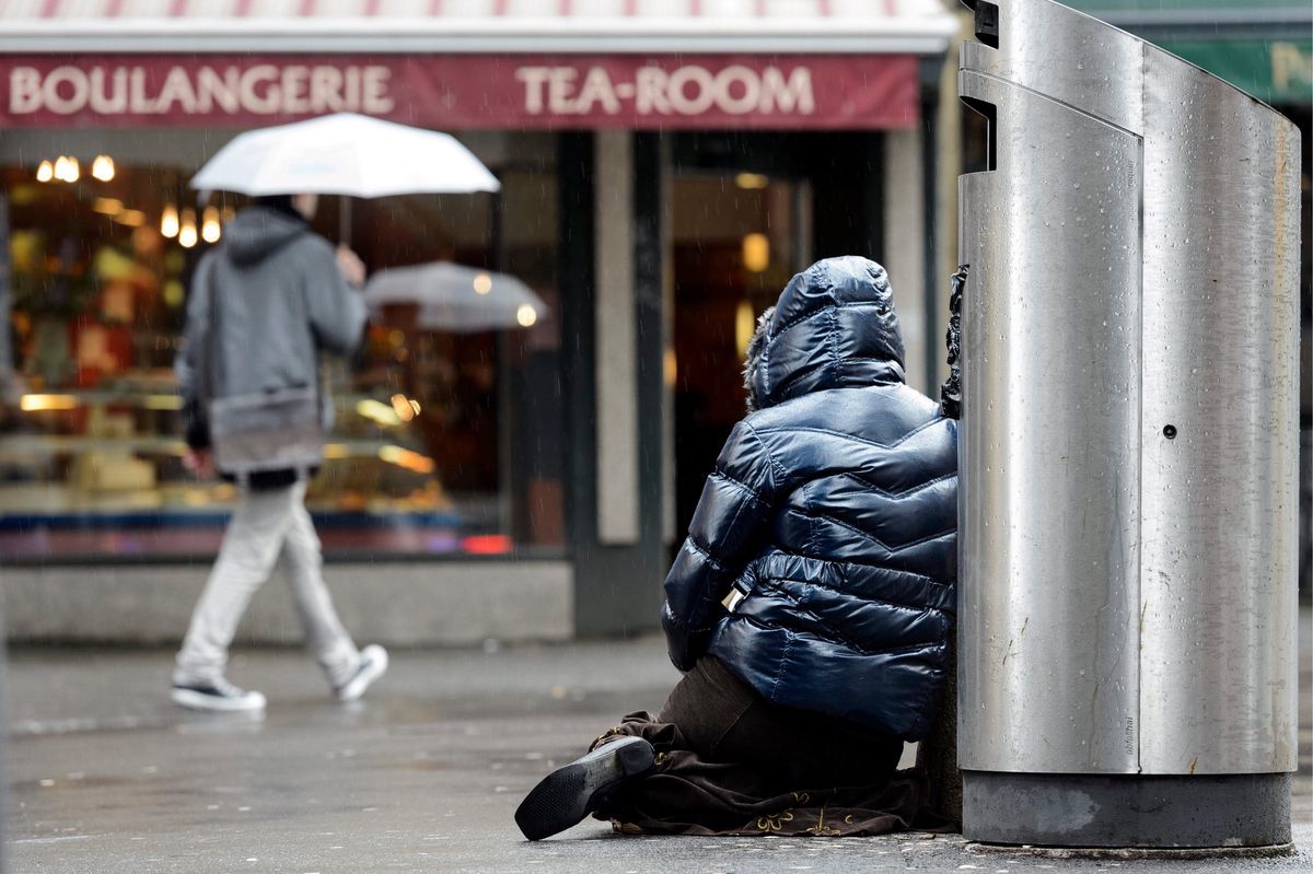 Un mendiant assis près d’une poubelle sur une rue piétonne sous la pluie à Lausanne, avec une boulangerie en arrière-plan.