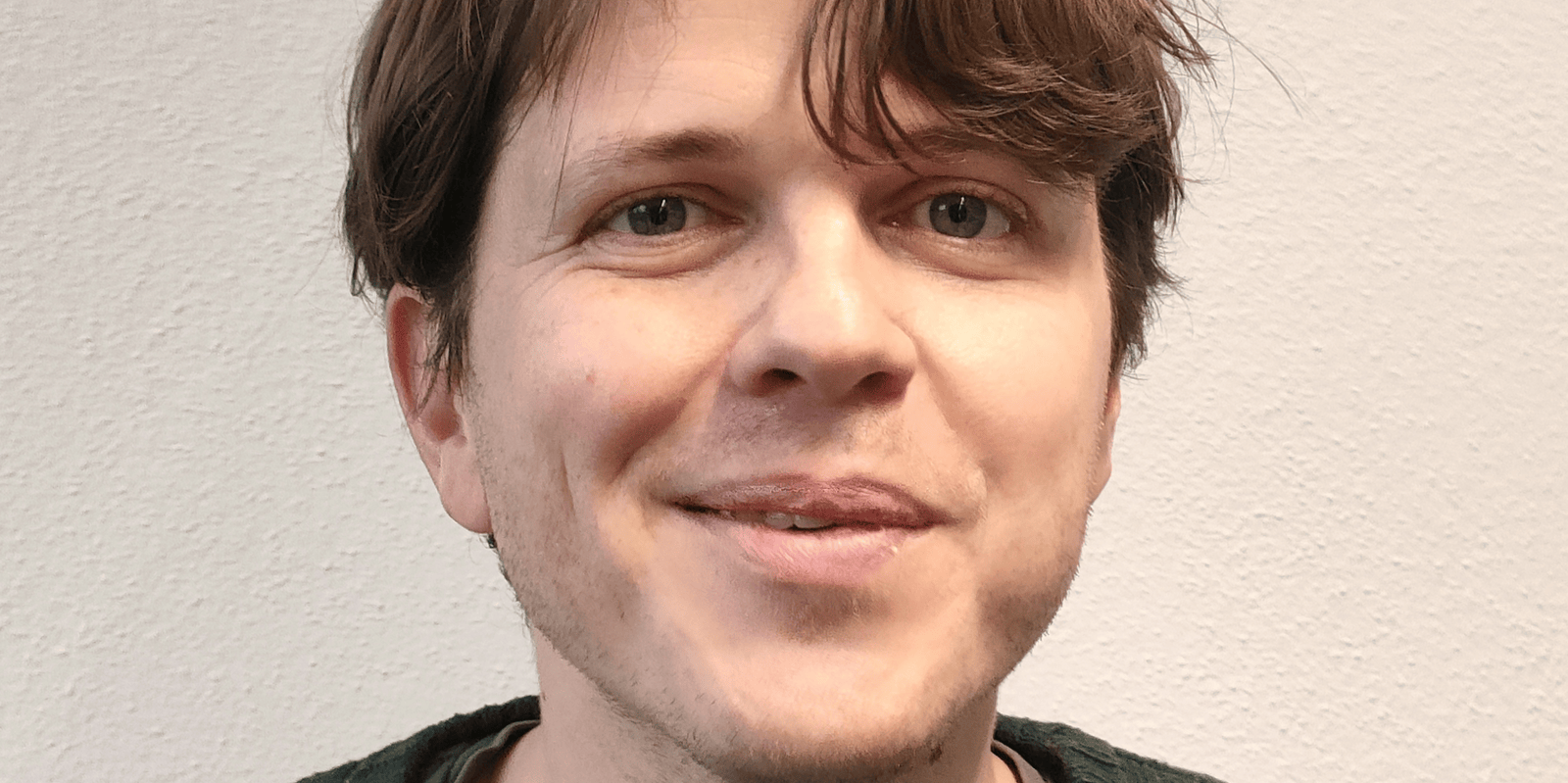 Personne souriante portant un pull vert devant un mur blanc.