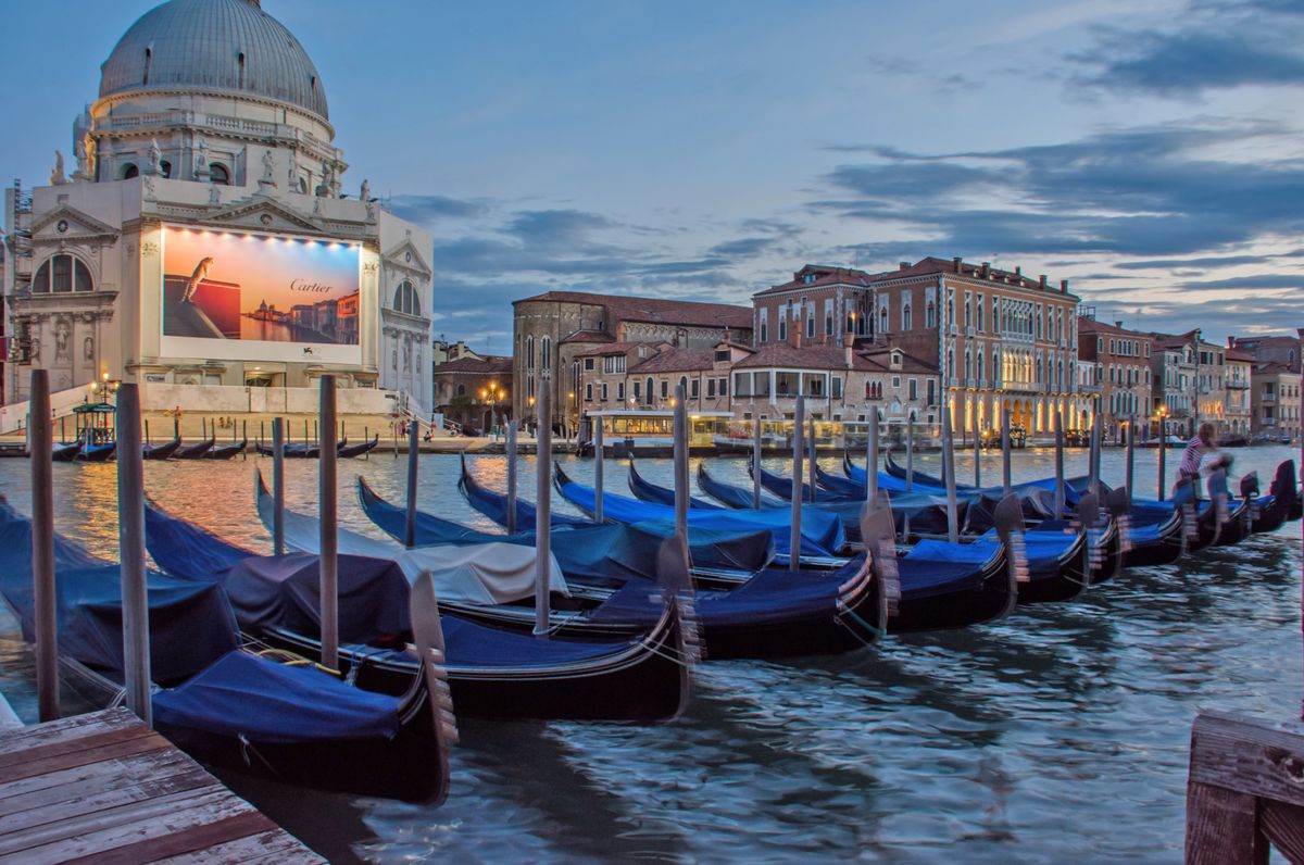 Une rangée de gondoles amarrées à un quai devant une église avec un dôme dans une scène de coucher de soleil à Venise, Italie.