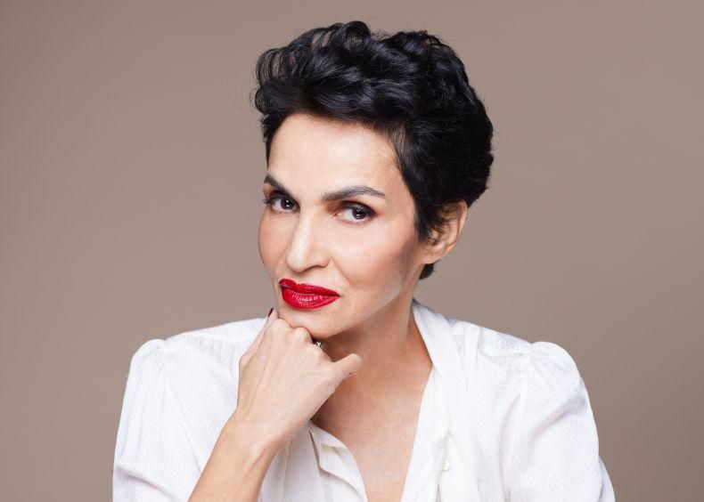 Femme aux cheveux courts noirs et lèvres rouges, portant une chemise blanche, posant avec la main sous le menton.