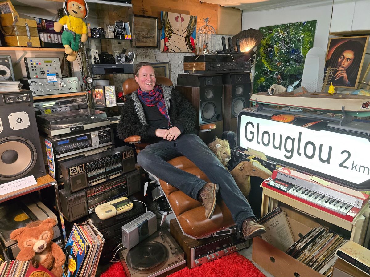 Homme détendu dans une pièce remplie de matériel audio vintage, avec une signalisation ’Glouglou 2km’ et des objets rétro.