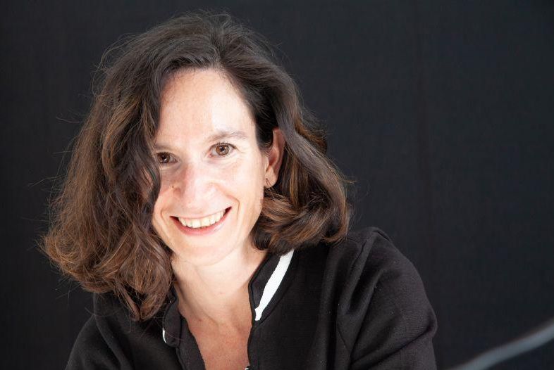 Une femme souriante aux cheveux bruns, portant un pull noir avec un col blanc, sur un fond noir.