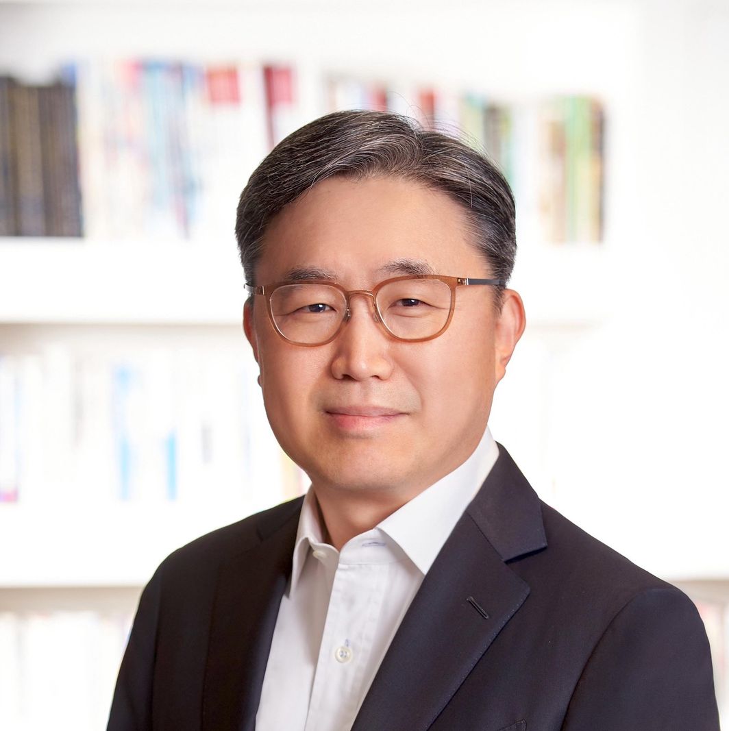 Porträt eines Mannes in einem dunklen Anzug mit Brille. Im Hintergrund sind unscharfe Bücherregale zu sehen.