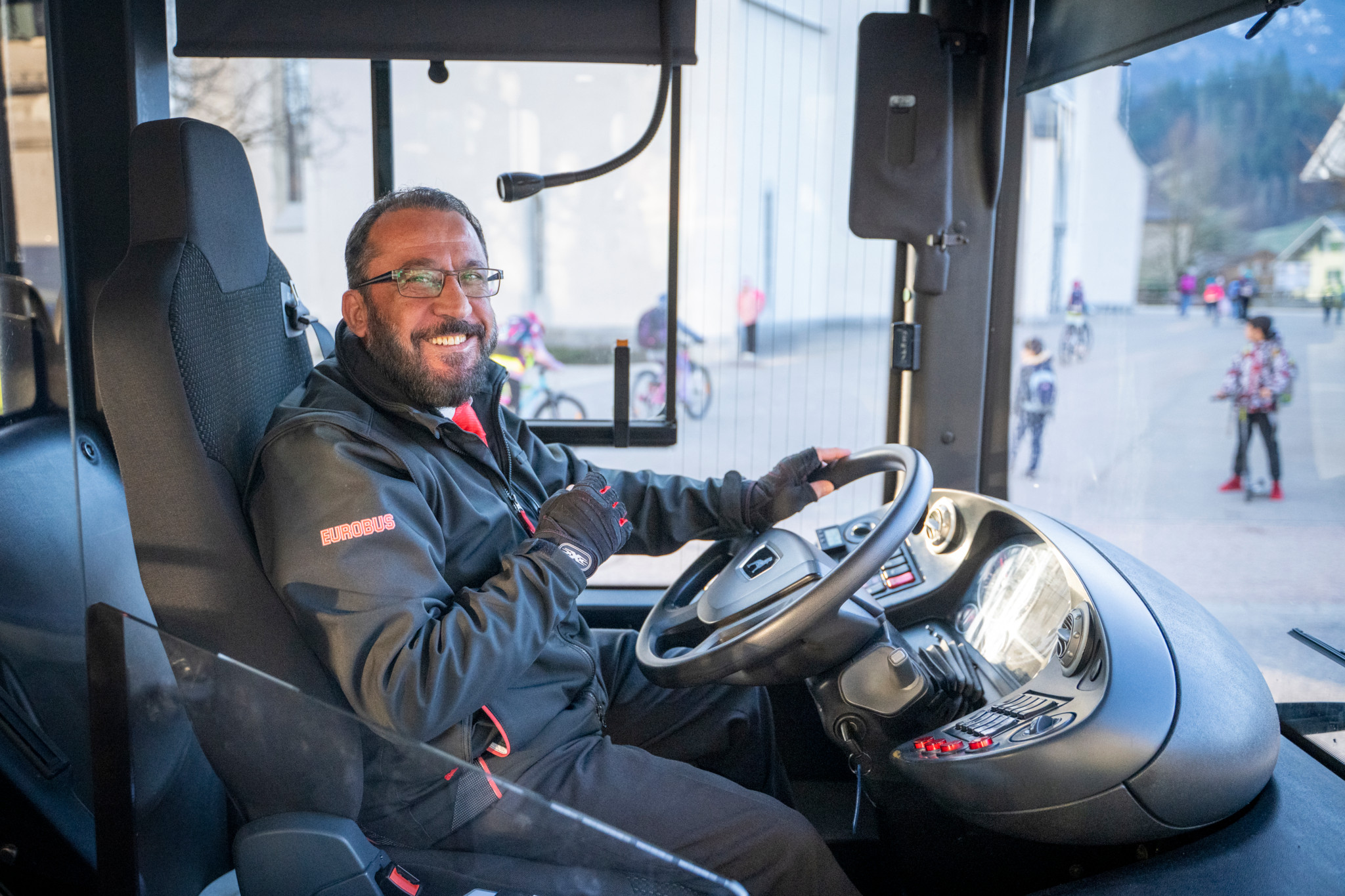 Busfahrer lächelt im Bus von Langnau nach Schüpfheim. Hintergrund zeigt Fahrgäste an einer Haltestelle. Foto: Marcel Bieri