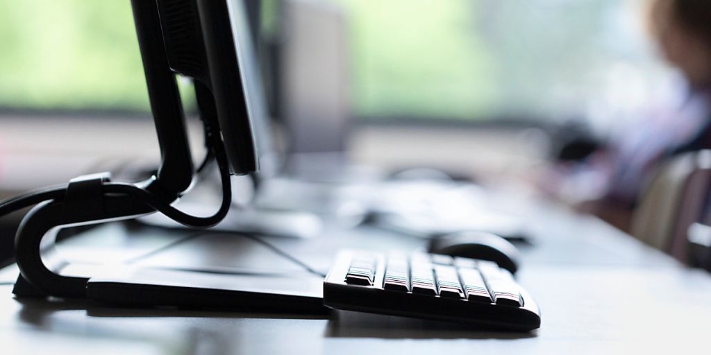 Seitenansicht eines Computerbildschirms und einer Tastatur in einem Büro. Unscharfer Hintergrund mit einer Person.