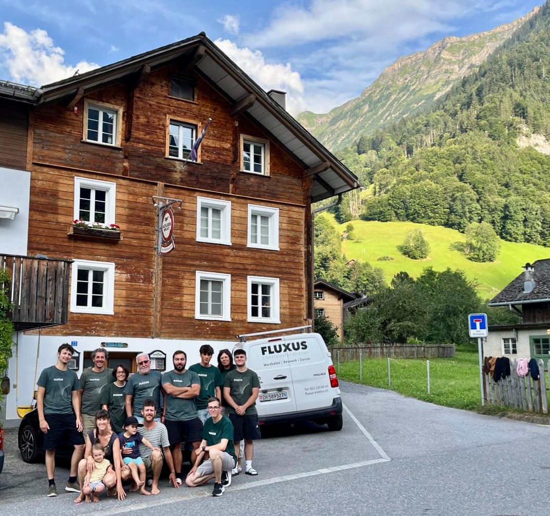 Eine Gruppe von Menschen posiert vor einem traditionellen Holzhaus mit einem weissen Van im Vordergrund. Im Hintergrund sind grüne Hügel und ein Berg zu sehen.