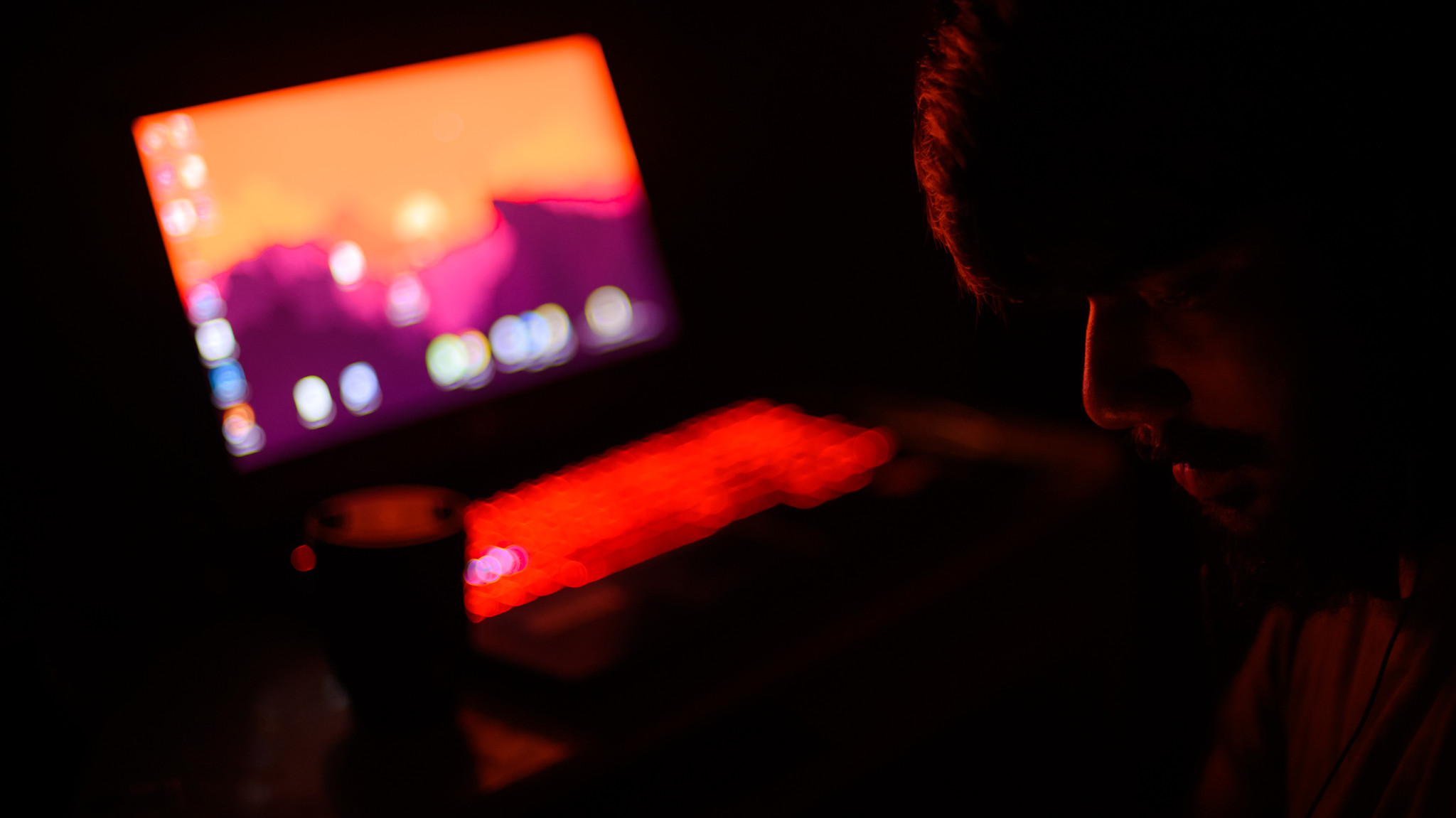 Person sitzt im Dunkeln vor einem Laptop mit leuchtendem rotem Keyboard und buntem Bildschirm. Person sitzt im Dunkeln vor einem Laptop mit leuchtendem rotem Keyboard und buntem Bildschirm.