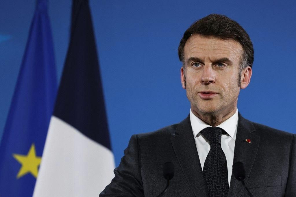 Homme en costume sombre parlant devant les drapeaux de l’UE et de la France sur un fond bleu.