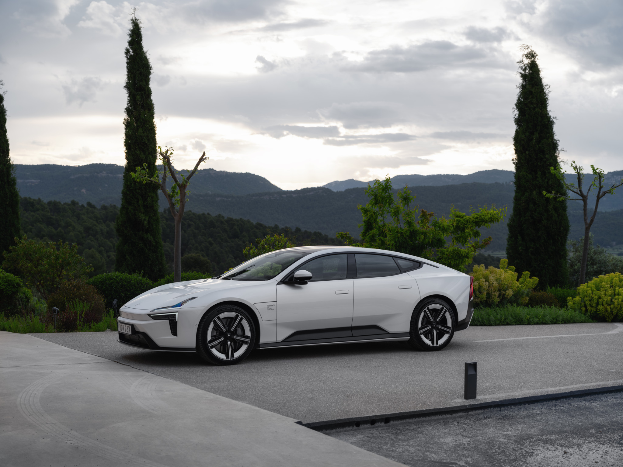 Weisses Elektroauto parkt vor einer mediterranen Landschaft mit Zypressen und Bergen im Hintergrund bei bewölktem Himmel.