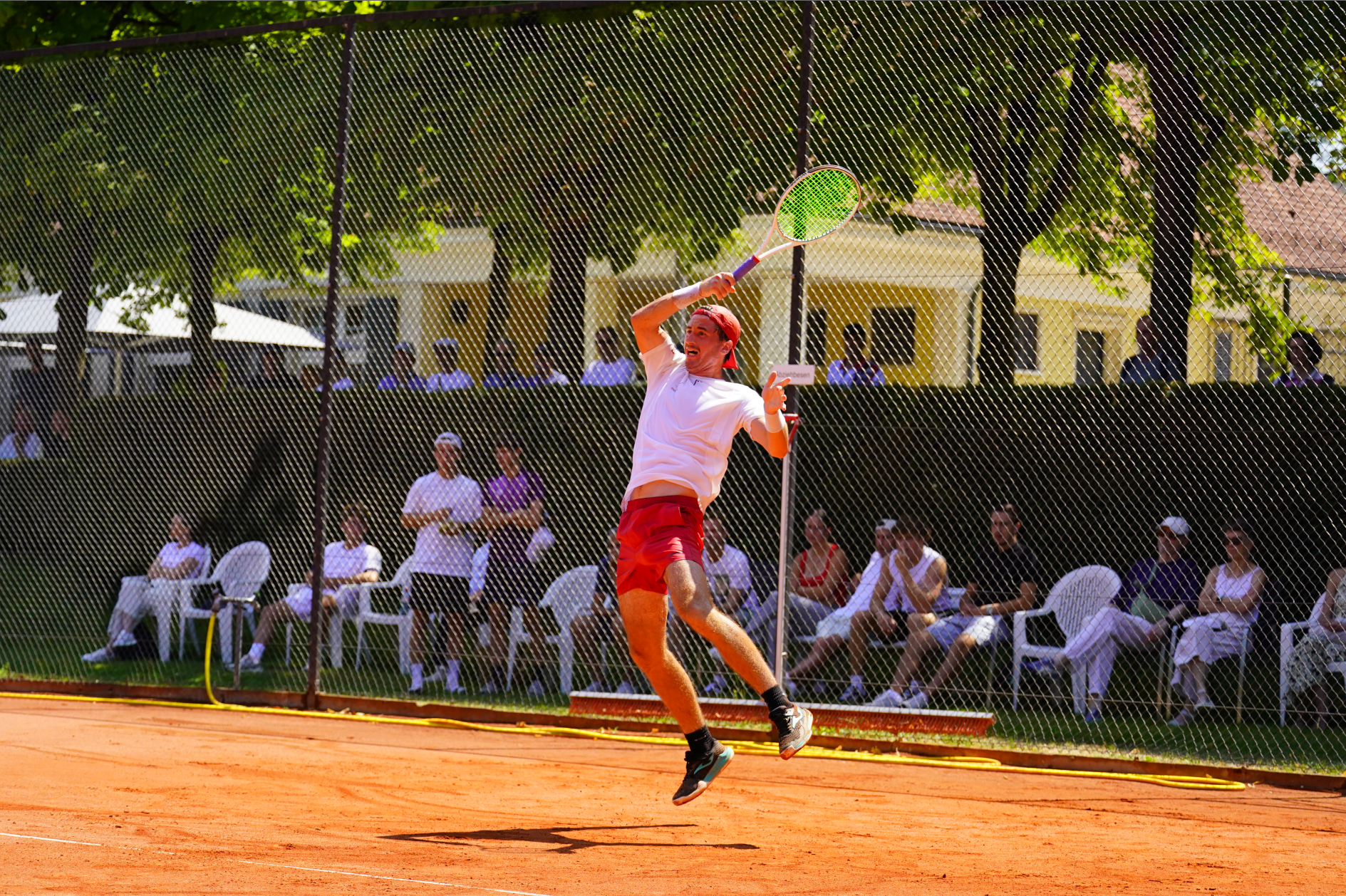Tennisspieler in roter Shorts springt für einen Schmetterball auf einem Sandplatz, Zuschauer im Hintergrund.