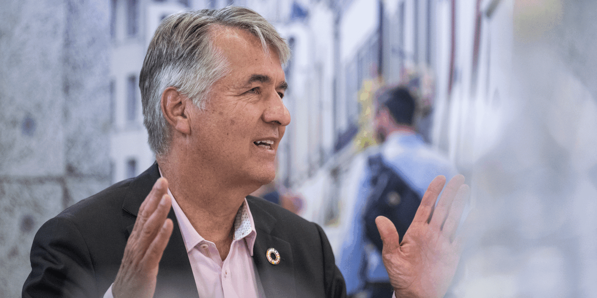 Ein Porträt von Alec von Graffenried. Er trägt einem Anzug gestikuliert, während er spricht; verschwommener Hintergrund mit Menschen im Freien.