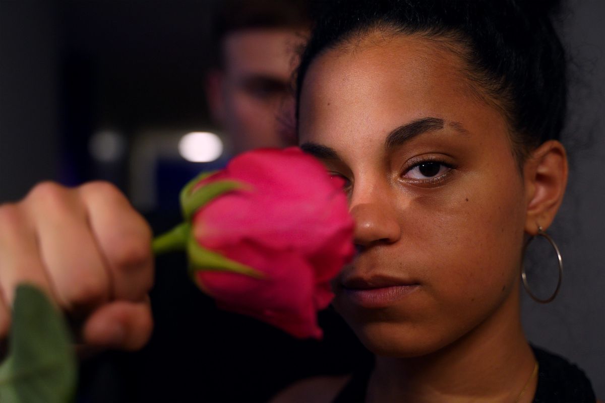 Une femme tenant une rose rose près de son visage, regardant fixement l’objectif, avec une personne floue en arrière-plan.