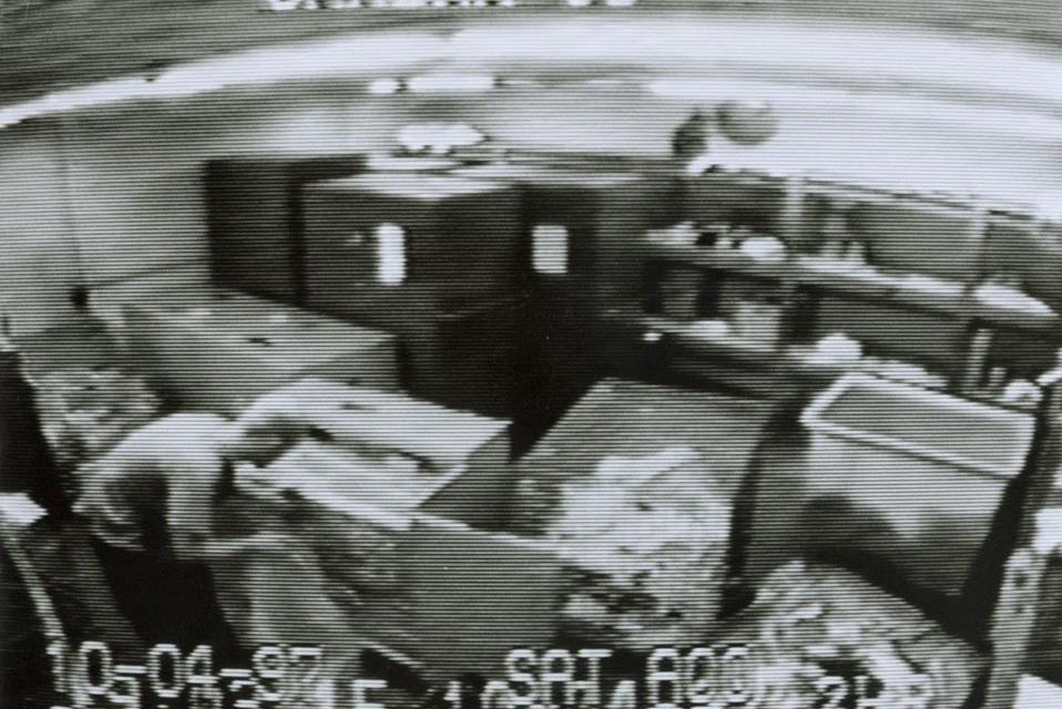 Image de sécurité en noir et blanc montrant l’intérieur d’une cuisine industrielle avec des équipements de cuisine et des étagères.