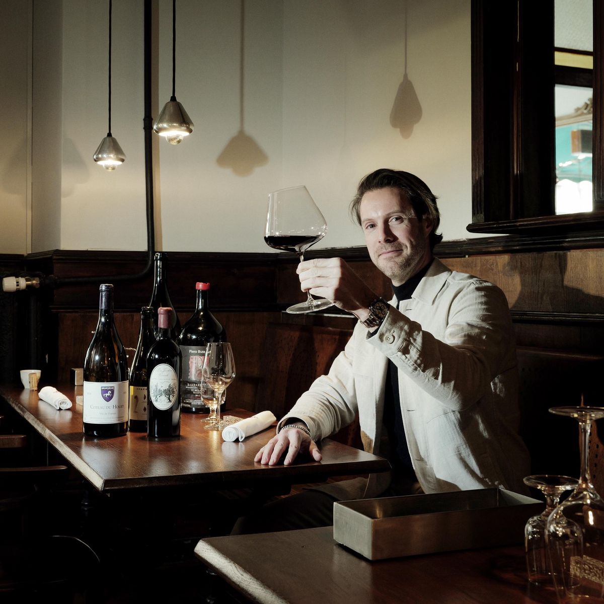 Pablo Nedelec, fondateur de Vins Vibrants à Genève, levant un verre de vin rouge, assis à une table avec diverses bouteilles de vin exposées.
