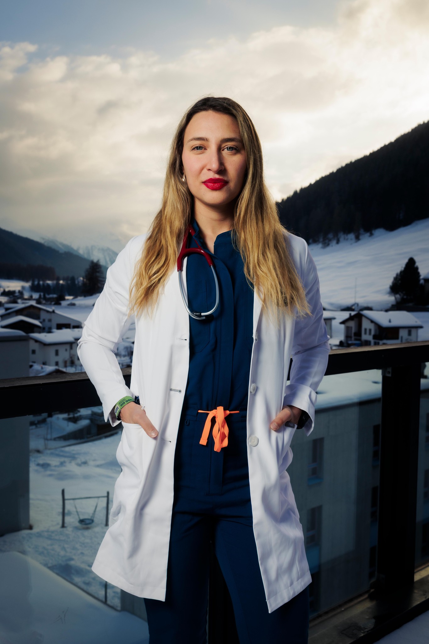 Ärztin im weissen Kittel steht auf einem Balkon mit schneebedeckten Bergen im Hintergrund.