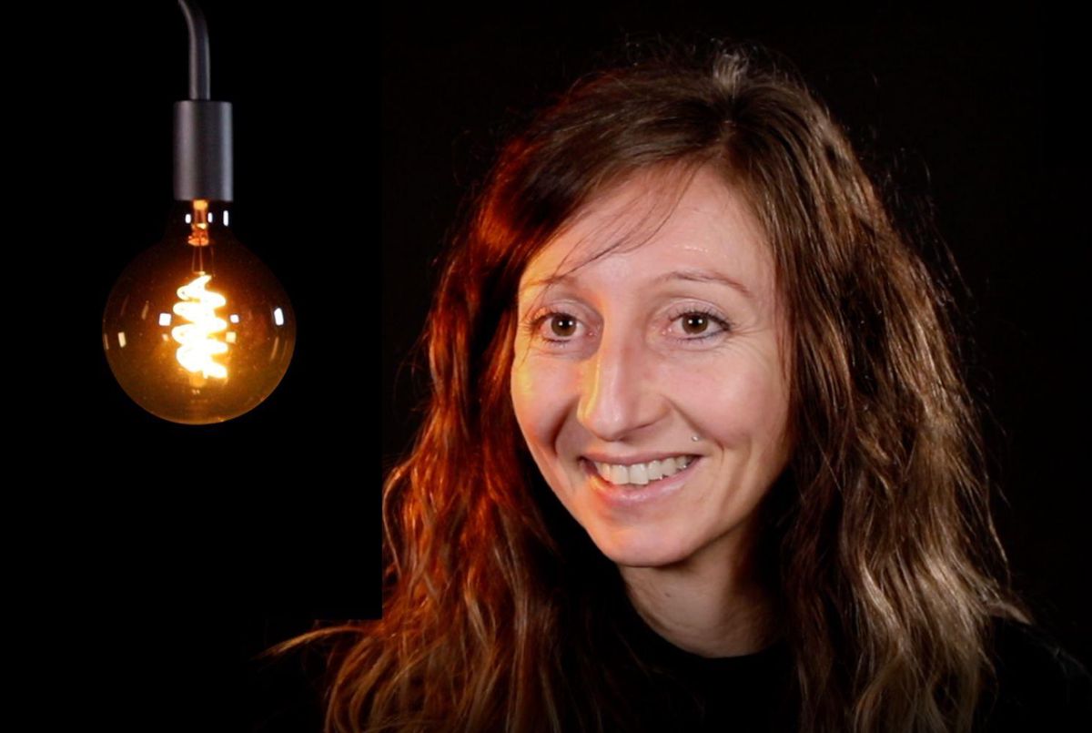 Femme souriante avec des cheveux bouclés se tenant près d’une ampoule suspendue allumée.