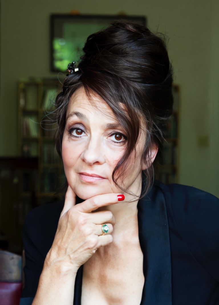 Femme aux cheveux coiffés en chignon, tenant son menton avec sa main, portant un vernis rouge et une veste noire.