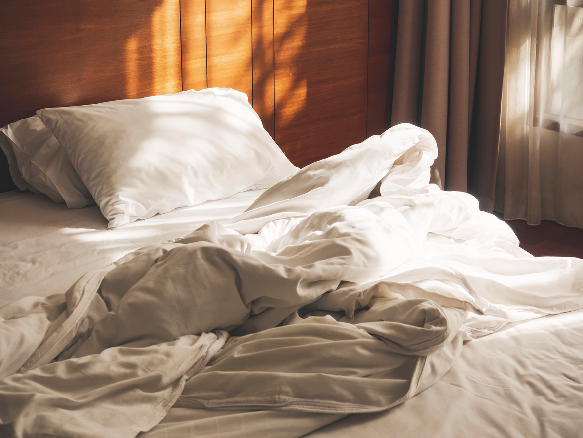 Lit défait le matin avec oreillers blancs, couette froissée et lumière du soleil filtrant à travers les rideaux Lit défait le matin avec oreillers blancs, couette froissée et lumière du soleil filtrant à travers les rideaux
